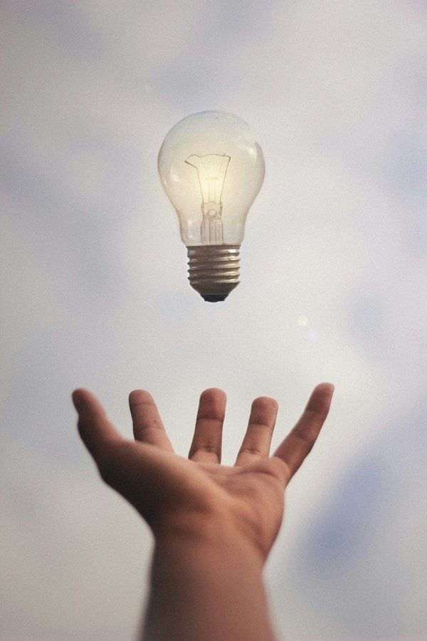 A hand reaching up toward a glowing light bulb against a cloudy sky